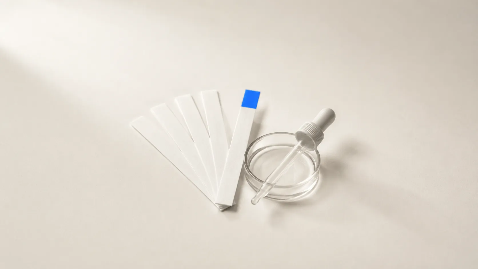 Fünf weisse Teststreifen fächerförmig auf beigem Untergrund, der vorderste mit blau eingefärbtem Feld, daneben eine flache Glasschale mit Pipette